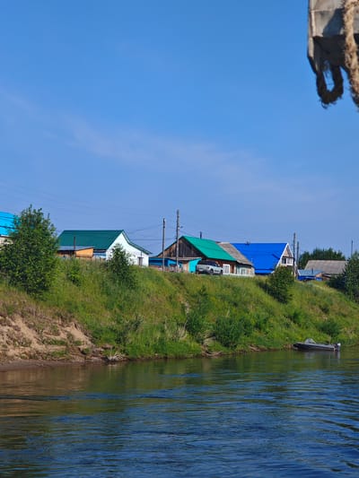 In Galanino, Russia, a serene river flows calmly under a clear blue sky. The riverbank, covered in lush green grass and dotted with trees, slopes upwards towards a row of houses. These houses, characterized by their colorful roofs in shades of green and blue, stand as a charming testament to rural architecture. A silver car is parked in front of one of the houses, suggesting a quiet, residential atmosphere. A small motorboat is tethered to the shore, ready for a leisurely ride on the water. The scene is peaceful and idyllic, evoking a sense of tranquility and the simple beauty of nature.