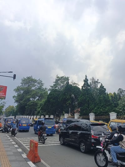 The image captures an overcast urban street scene in the city of Soibada, East Timor, depicting moderate to heavy traffic. The sky is largely obscured by dense, light grey cloud cover. In the background, a large area of dark green trees, including several tall pine trees and dense foliage, rises above a decorative fence featuring white pillars and dark metal railings.
The roadway, constructed of asphalt, is visible in the lower half of the frame and includes a prominent white zebra crossing. On the left side, a brown-tiled sidewalk is present, with an orange plastic traffic barrier placed in the foreground. Overhead, a black traffic light is suspended from a pole on the far left, beneath a partially visible red banner.
The traffic flow consists of numerous vehicles. Several blue vans, possibly public transportation, are observed, with one positioned over the zebra crossing. A black SUV, consistent with a Toyota Avanza model, is visible in the right lane, facing away from the camera. Portions of yellow vehicles are present further back on the right. Multiple motorcycles are interspersed throughout the lanes, with riders wearing helmets. One individual on a dark motorcycle is positioned on the zebra crossing, moving right. Another individual, with a passenger, is on a white and pink motorcycle in the extreme right foreground, also facing away from the camera. The scene suggests an active urban intersection or roadway experiencing congestion.