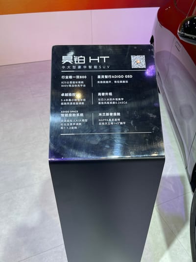 The image shows a black sign with white Chinese characters and English text. The sign is located at the Hangzhou International Expo Center, Hangzhou, China (latitude: 30.272, longitude: 120.416), where an automotive expo is taking place. The sign is attached to a black pedestal and sits next to a red car. The sign displays the features of the “Realme HT,” a Chinese brand of electric car. Features include a range of 800 km, 825 mm of legroom, 5.6 mm of ground clearance, and a 143-square-meter space. The sign also advertises a QR code that leads to further information about the car.