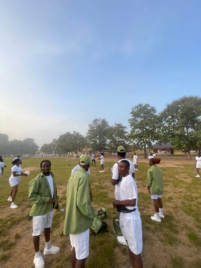 In Emure-Ekiti, Nigeria, a group of young adults, likely participants in a National Youth Service Corps (NYSC) program, are gathered in an open field under a hazy sky. The scene suggests an outdoor activity, possibly a sporting event or training session, as several individuals are holding or interacting with a volleyball.

The majority of the individuals are dressed in white t-shirts and shorts, with some wearing green jackets or shirts over their white attire, consistent with typical NYSC camp uniforms. A few people are wearing green caps. The environment is a grassy field with scattered trees in the background, and a building with a yellow facade is visible further away. The lighting indicates it is either early morning or late afternoon due to the soft light and haze.

Focusing on the foreground, a young man in a green jacket and white shorts is looking towards the camera, with another person in a similar outfit partially visible to his right. To the left, a woman in a white t-shirt and shorts is holding a volleyball. Other individuals are dispersed across the field, some engaged in conversation or preparing for activities. The overall atmosphere appears relaxed and communal.