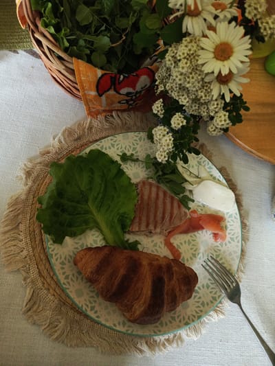 A close-up, high-angle, overhead shot shows a plate of food set on a textured placemat and a white tablecloth. The plate, which has a light blue and white patterned rim, contains a croissant, a large green lettuce leaf, a piece of grilled meat, some thinly sliced pink fish, a dollop of white cheese, and a few sprigs of green herbs. A silver fork rests to the right of the plate. In the background, a woven basket filled with greenery is visible, along with a bouquet of white flowers and daisies. The overall impression is of a prepared meal. The location is identified as Alexandrovskaya, Russia, although no specific visual cues in the image confirm this.