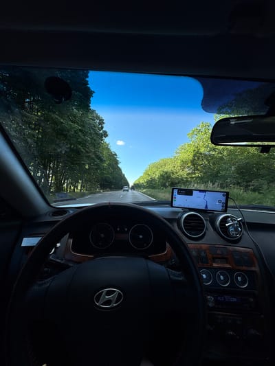 The image captures a point-of-view shot from the driver's seat of a car, looking forward through the windshield. The car is on a road lined with dense green trees on both sides, creating a canopy effect. The sky is a clear, bright blue with a few wispy clouds.
In the foreground, the steering wheel of a Hyundai is visible, featuring red stitching. The dashboard displays a navigation screen on a smartphone mounted centrally. The screen shows a map with route guidance, indicating 0.6 km remaining to a destination. To the right of the navigation device are air vents and control buttons. The rearview mirror is visible at the top right of the frame.
Further down the road, two cars are visible in the distance, traveling in the same direction. The environment suggests a rural or suburban setting, likely a scenic drive on a sunny day. The location is specified as the city of Stefanovo, Bulgaria.