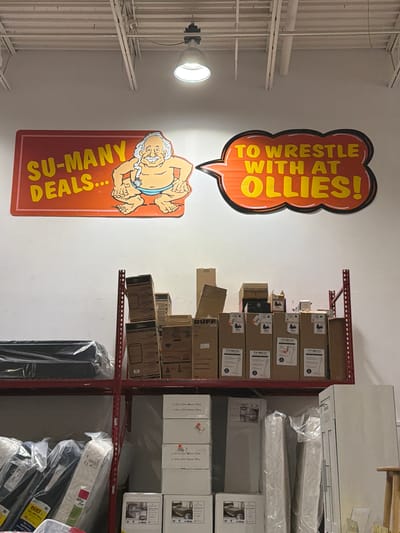 A photograph taken at an indoor retail establishment, possibly a discount store, features a large, brightly colored sign on a white wall. The sign, split into two sections, reads "SU-MANY DEALS..." on the left, accompanied by a cartoon illustration of a jovial, overweight man in a blue loincloth. The right section of the sign, in a speech bubble shape, states "TO WRESTLE WITH AT OLLIES!". The background above the sign shows industrial ceiling fixtures and lighting. Below the sign, metal shelving units are filled with various boxed goods. These boxes, some labeled with brands like "TINECO" and "DEHUMIDIFIER," are stacked on the shelves. In the foreground and lower sections of the image, more merchandise is visible, including items wrapped in plastic sheeting, possibly mattresses or furniture. The overall scene depicts a retail environment stocked with goods, advertising a sale or promotion with a playful mascot. The specific location of Elizabeth City, United States of America, is not explicitly indicated by any visual cues within the image itself.
