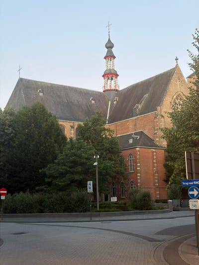The image depicts the historic Saint Peter's Church (Sint-Pieterskerk) in Turnhout, Belgium, viewed from the street. The church is a prominent brick structure with a distinctive red and white bell tower topped with a cross. The main building features a large, dark, sloping roof and arched windows. Surrounding the church are mature trees and green bushes. In the foreground, a paved street curves to the right. Traffic signs are visible: a red and white circular sign indicating "no entry" or "prohibited," and a blue directional sign with text in Dutch ("Terug naar RING") indicating a return route to the ring road, along with an arrow pointing right and a bicycle symbol indicating an exception. The sky is clear and pale blue.