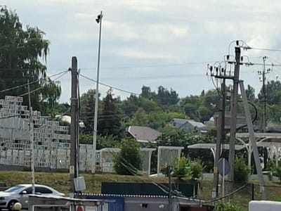 The image depicts an outdoor scene in Verkhny Uslon, Russia, likely during the daytime. The sky is overcast, suggesting cloudy weather.
The foreground shows a white car parked on the left, partially visible. In front of the car and extending to the right is a low structure, possibly a kiosk or a small building, with some roofing and signage that is not clearly legible.
Behind the foreground structures, a sloped grassy area rises. A white fence runs along this embankment, with some sections featuring vertical bars and others appearing more solid. Beyond the fence, there are numerous trees and greenery, indicating a suburban or semi-rural setting. Several residential buildings with grey roofs are visible nestled amongst the trees in the middle ground.
Dominating the right side of the image are utility poles with electrical infrastructure, including insulators and wires. A tall, thin pole with what appears to be two lights or cameras is positioned towards the left of the image, also featuring overhead wires.
In the far left, a stack of white, crate-like structures is visible, possibly storage or temporary structures. The overall scene appears to be a relatively ordinary day with no obvious events or activities taking place. The image has a slightly blurred quality, making precise details difficult to discern.