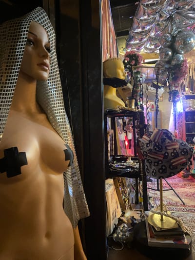 The image depicts a storefront display in Baan Nai Suan, Thailand. A mannequin, partially nude with black crosses covering the nipples and a silver checkered head covering, lies horizontally in the foreground. Behind the prone mannequin, there is a yellow mannequin head and a display rack containing jewelry, a hat with a star and stripe pattern, and various other items. In the background, there are reflective disco balls and floral arrangements. The overall scene appears cluttered and eclectic, typical of a retail environment.