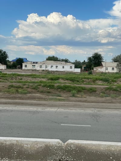 A street view in Qtay, Uzbekistan, captures a cloudy day with a vibrant blue sky. The foreground features a concrete barrier and asphalt road with a visible white line. Beyond the road, a strip of green vegetation leads to a collection of buildings. These structures appear to be single-story homes, some with white walls and tiled roofs. Solar panels are visible on the roof of one building. Trees dot the landscape, and a blue-tiled roof of another building is noticeable. The scene depicts a residential area with signs of wear and tear on some of the buildings.