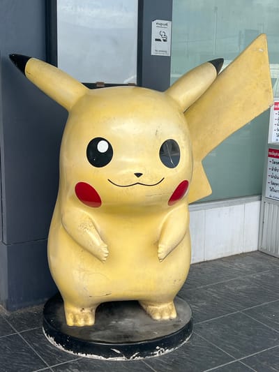 An outdoor, eye-level, full shot shows a large, weathered, yellow Pikachu statue. The statue stands on a black circular base, which is chipped and worn around the edges. The Pikachu statue is slightly dirty and has some cracks on its face and body. Its eyes are black with white pupils. It has red circles on its cheeks and a small black mouth. The statue has two pointed ears, one with a black tip, and a lightning bolt-shaped tail. In the background, there is a building with a dark blue wall and large windows. A "No Smoking" sign is visible on the left side of the building. To the right of the statue, there is a white wall and a metal gate. The ground is tiled with dark gray squares. The overall scene is located in Ban Ko Pru, Thailand.