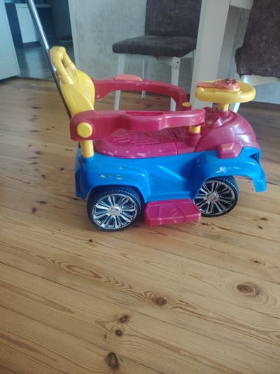 A toy car with a push handle, steering wheel, and seating is displayed on a wooden floor. The car is primarily red, blue, and yellow. In the background, a chair with a dark upholstery is visible, along with part of a wall. The environment appears to be indoors. The image is taken from a slightly elevated perspective, looking down at the toy car. The floorboards are light brown with visible wood grain and knots. The overall scene is well-lit.