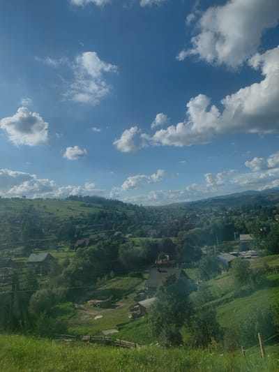 The image displays a panoramic, elevated view of Lazeshchyna, Ukraine, characterized by rolling green hills under a bright blue sky with numerous scattered white cumulus clouds. The landscape consists of undulating terrain heavily vegetated with green grass and dense clusters of deciduous and coniferous trees, extending to hazy, forested mountains in the background. A rural settlement is dispersed across the lower slopes and valley floor, featuring numerous residential structures, primarily single-story, with dark-colored roofs and lighter walls. Unpaved dirt paths connect some of these buildings; one such path in the mid-ground contains a large, light-colored vehicle, likely a truck or agricultural machinery, positioned near a distinct circular ground feature. A rustic wooden fence runs horizontally across the lower foreground, and a dark utility pole with wires is visible on the left-central side of the frame. No individuals, specific actions, or interactions are discernible.