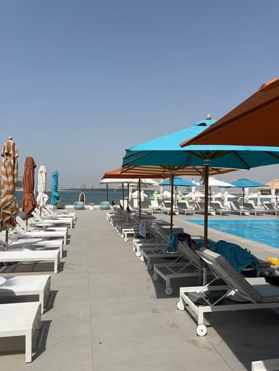 This is a medium shot, taken outdoors, of a resort pool area with a beach and the ocean in the background. The scene is likely during the day due to the bright sunlight casting shadows. The sky is clear and blue, suggesting fair weather.
In the foreground and midground, there are many white lounge chairs and a row of closed and open umbrellas. The umbrellas vary in color, with some being striped brown and tan, others solid blue, and one large orange umbrella is partially visible. Several blue towels are draped over the lounge chairs. The lounge chairs are arranged on a light gray tiled surface, with a swimming pool visible to the left. The pool water is blue and appears calm.
In the background, the ocean stretches out, with a distant cityscape and construction cranes visible on the horizon. This suggests an urban or developing coastal location. There are no people clearly visible in the image. The overall mood of the scene is peaceful and suggests a relaxing vacation or resort atmosphere. The location is identified as Ras Al-Khaimah, United Arab Emirates.