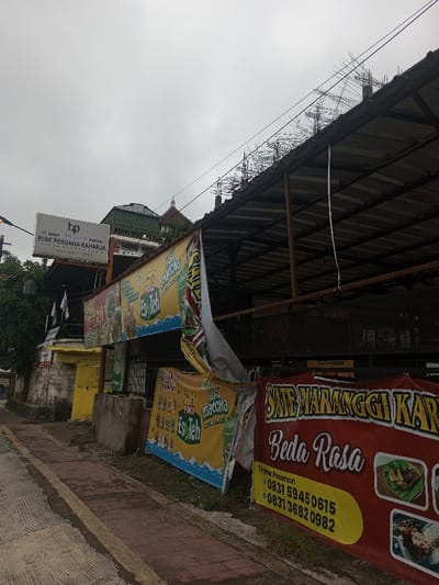 The image depicts a street scene on an overcast day, featuring several buildings and numerous commercial banners.
In the foreground, a brick-patterned sidewalk with a yellow painted line runs along the bottom left. To the right of the sidewalk, several large, colorful banners are prominently displayed. The most dominant banner in the lower right corner is red with yellow text, advertising "SATE MARANGGI KAR Beda Rasa" (Maranggi Satay Different Taste). This banner also includes images of prepared food dishes and contact information for orders: "Terima Pesanan 0831 5945 0615" and "0831 3682 0982".
Behind and to the left of the red banner, multiple yellow banners with green and blue accents are hung, advertising "POCI Es Teh" (Poci Iced Tea) and "Maccha" (Matcha), featuring images of cold beverages. One of these yellow banners is partially torn and hanging loose in the middle of the frame. A gray concrete block or planter sits on the ground near these banners.
Above the banners, an open-sided structure with a dark corrugated roof is visible. This structure appears to be in an unfinished or partially dilapidated state, with numerous pieces of rebar protruding upwards along the roofline, suggesting ongoing construction or a building that has seen better days. Overhead, several power lines or communication cables stretch across the top portion of the frame.
In the mid-ground on the left, a white signboard is mounted on a pole or building. It displays the logo "bp" and the text "PT BANK PEREKONOVAN RAKYAT BUMI PENDAWA RAHARJA (Sejak Tahun 1990)," indicating a bank established in 1990. Behind this sign and the yellow banners, a green-roofed building is partially visible, along with a small, dome-like structure, possibly the dome of a mosque. Further to the left, lush green foliage is present.
The sky is uniformly gray and overcast, suggesting a cloudy day with low light conditions. The overall impression is a bustling commercial area, likely in an Indonesian town or city, characterized by roadside advertising and visible infrastructure.