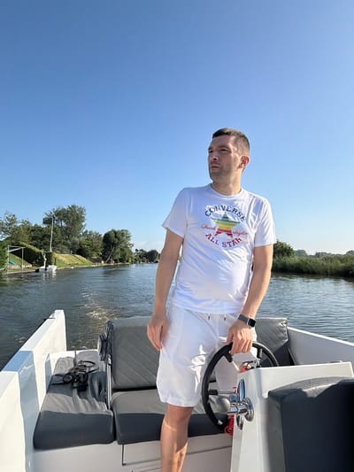 In Rhenoy, Netherlands, a man stands at the helm of a white motorboat on a calm waterway under a clear blue sky. He is wearing a white t-shirt with a colorful Converse All Star logo and white shorts. His left hand is on the steering wheel, and his right hand is resting on his hip. The boat's interior features gray upholstered seats. The surrounding environment consists of lush green trees and vegetation lining the water's edge, with a few other boats visible in the distance. The wake behind the boat indicates it is in motion.