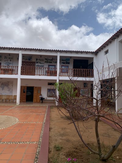 The image shows the courtyard of a building in Boca del Río, Venezuela. The courtyard is paved with terracotta tiles and surrounded by a white, two-story building with a red tiled roof. The building has open balconies on the upper floor, adorned with wooden railings. Various framed pictures and maps are displayed on the walls of the building. In the foreground, a bare, thorny tree stands in the sandy ground. A single person is seated on a bench to the right of an open doorway, looking at a device in their hands. Several wooden chairs are arranged in a row along the left side of the courtyard. The sky above is partly cloudy.