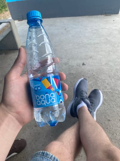 A person's hand holds a clear plastic bottle of Bona Aqua water. The bottle has a blue cap and a label with colorful geometric shapes and the text "bona aqua" and "НЕГАЗИРОВАННАЯ" (non-carbonated). The bottle appears partially full, with condensation on the outside. The person is wearing a blue denim garment, visible at the bottom of the frame, and grey athletic shoes with white socks. They are seated on a grey concrete surface, with their legs crossed. In the background, there is a dark grey or blue structure and some greenery. The scene is outdoors, possibly in an urban or recreational area.