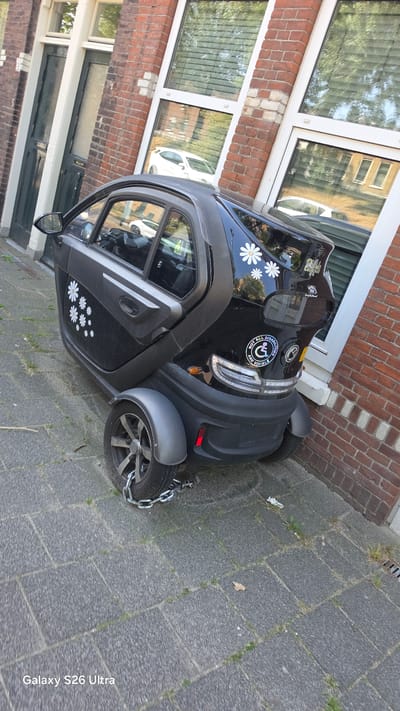 This image shows a small, black, three-wheeled electric vehicle parked on a paved sidewalk in front of a brick building. The vehicle has a single door on the left side and a roof. White daisy stickers are visible on the side door and the rear of the vehicle. A round sticker with text is also present on the rear, reading "ALL DISABILITIES ARE NOT VISIBLE". A metal chain is wrapped around the rear wheel, likely for security. The building has large windows reflecting trees and other parked cars. The ground is paved with grey square tiles, with some grass growing between them. The scene appears to be outdoors during the daytime, with bright lighting suggesting sunny weather. The presence of the vehicle, building, and paved sidewalk indicate an urban or suburban residential setting in Vlaardingen, Netherlands. There are no people visible in the image.