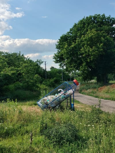 The image depicts an outdoor scene with a blue wire mesh recycling bin, shaped like a rocket, filled with plastic bottles and other waste. The bin is tilted and situated in tall, overgrown grass next to a gravel path. In the background, there are lush green trees, a utility pole with wires, and a clear blue sky with scattered white clouds. The location is identified as Lykhove, Ukraine. There are no people visible in the image. The overall impression is one of a rural or semi-rural setting with a focus on waste disposal in a natural environment.