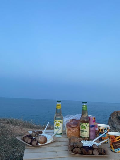 This media file shows a close-up view of food and drinks arranged on a wooden surface, with the sea and a rocky cliff in the background. The scene appears to be a picnic or an outdoor meal.
The subjects include two bottles of Somersby cider (one pear-flavored and one apple-flavored), two disposable coffee cups with colorful designs featuring coffee-related imagery and text like "CAPPUCCINO" and "Espresso," and a can of "SOLUT DRINKS RASPBERRY MONADE." A paper tray holds a portion of food consisting of several pieces of grilled meat, likely sausages and chicken, served with a dollop of white sauce or cream. A plastic-wrapped bread roll is also visible. Two plastic cutlery pieces are in the food tray.
The setting is outdoors, likely near a coast, given the view of the sea and the rocky terrain with sparse vegetation. The overall lighting suggests it might be late afternoon or early evening, with a soft, diffused light. The sky is a clear, pale blue, indicating fair weather.
The visible text on the items includes brand names like "SOMERSBY," "SOLUT DRINKS," and descriptions of the flavors like "APPLE," "PEAR," and "RASPBERRY MONADE." The coffee cups also have text related to coffee drinks.
There are no people visible in the frame, but the arrangement of food and drinks implies a gathering or a personal treat. The overall mood seems relaxed and leisurely, suitable for enjoying a scenic view with refreshments. The location context provided, Tyulenovo, Bulgaria, suggests a coastal setting in that region.