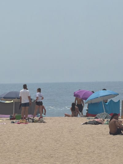 This is a photograph of a sandy beach in Espinho, Portugal, on a clear, hazy day. The ocean is visible in the background, with a calm sea. In the foreground, several people are present, some under beach umbrellas.
On the left side of the frame, two individuals are standing near a dark blue umbrella and a gray beach tent. One person, wearing a white t-shirt and blue patterned shorts, is facing away from the camera. Beside them, a person in a white t-shirt and black shorts is also facing away. Scattered on the sand near them are various items, including a green bag, towels, and shoes.
Further to the right, under a bright purple umbrella, a person in black shorts is visible from the back. A blue beach umbrella is set up to the right of the purple one, with a blue and red beach tent behind it. Under the blue umbrella, a person wearing a bikini is lying down. Another person, also in swimwear, is seated to the left of the blue umbrella, facing the ocean.
In the mid-ground, further down the beach, a person in a black bikini is lying on a towel. To the far right, a person with long dark hair, wearing black shorts, is seated and looking to their left. A few beach towels and bags are spread out on the sand. The overall atmosphere suggests a relaxed day at the beach.