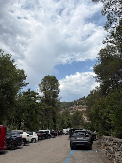 The image depicts a parking area in Montanejos, Spain. A row of parked cars lines the right side of the frame, with a gray Opel Mokka SUV closest to the viewer, displaying the license plate "4437 MVT". Further down the row are cars of varying colors including white and red. People are visible between the parked cars, some appearing to be standing or milling about.
On the left side of the image, dense green trees frame the scene. In the distance, a rocky cliff face rises above the trees. The sky is visible through gaps in the trees, showing white clouds and patches of blue sky.