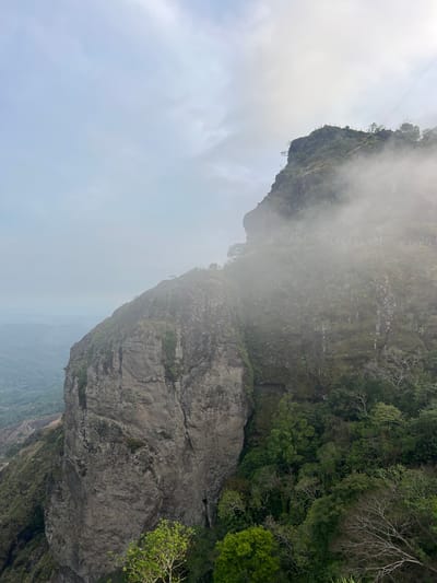 The image depicts a cliffside landscape in Planes de Renderos, El Salvador. The scene includes a prominent rock formation with vegetation clinging to its surface. Mist partially obscures the view, adding an element of atmospheric haze. The sky is overcast. A cable runs along the top. The overall impression is one of a natural, somewhat rugged environment.