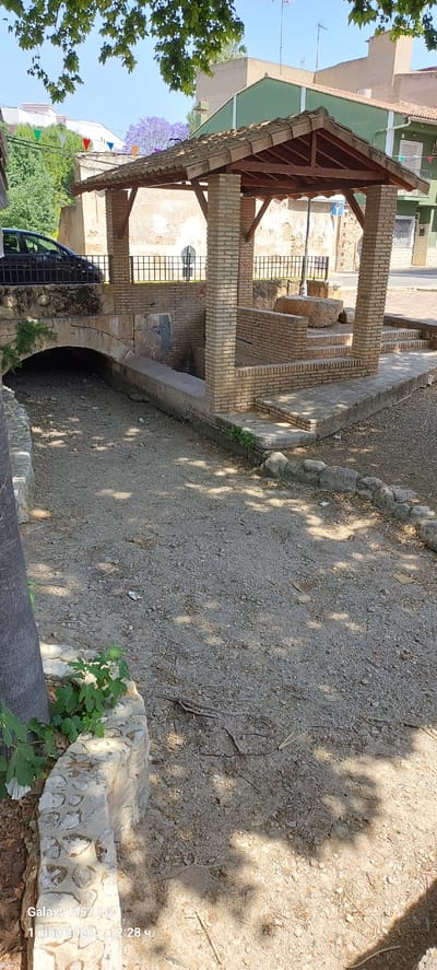 The image shows a partially shaded, gravel-covered ground in what appears to be a public space or park in the city of Canals, Spain. There is a brick structure with a roof, possibly a shelter or kiosk. The structure has an arched opening at the base. The area is enclosed by a stone wall, part of which is decorated with circular patterns. There is greenery, including trees and plants, visible. Buildings are visible in the background, suggesting the area is located within an urban or residential zone. A parked car is visible behind the brick structure. There is also some type of decoration such as pennants hanging overhead.