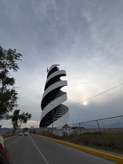 The image, correctly oriented, depicts a striking modern building with a distinctive spiral or wave-like facade, rendered in alternating white and dark grey/black bands. This architectural marvel stands under a mostly overcast sky, through which the sun casts a diffused, bright light from the upper right. A thin cable or wire extends horizontally across the sky, passing near the sun.

In the foreground, the tops of several dark green trees are visible on the left side of the frame. To the right of the building, a paved road stretches into the distance, featuring a prominent yellow curb line and white dashed lane markings. Several cars are discernible on the road, suggesting active traffic. Alongside the road, a chain-link fence separates it from a strip of wild, grassy terrain. In the background, distant mountains or hills are faintly visible, partially obscured by the atmospheric conditions.

The scene captures a mix of modern urban development and natural landscape, likely in an area like Tlaxcalancingo, Mexico, known for its blend of infrastructure and scenic backdrops. The time of day appears to be daytime, possibly mid-morning or late afternoon, given the diffused sunlight. The weather is cloudy but bright. There are no identifiable people or explicit activities beyond vehicles traveling on the road. No visible text is present in the image.