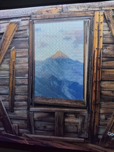 The image shows a close-up of a rustic wooden structure with a window. Through the window, a mountain landscape is visible, with a peak in the center under a cloudy sky. The wooden walls appear weathered and are constructed from horizontal planks. Diagonal wooden supports are attached to the exterior. The bottom right corner of the image shows text that appears to be a watermark or logo, with the word "Goo" visible. There are no people or animals in the image. The scene is not identifiable as being in Reftinsky, Russia.
