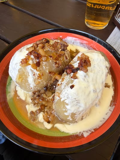 The image shows a close-up of a dish of cepelinai, a traditional Lithuanian potato dumpling, served in a colorful, striped bowl. The cepelinai are generously topped with a creamy sauce and crispy fried bacon bits. The dish is placed on a wooden table, and a glass of beer is visible in the background. The overall impression is one of a hearty, traditional meal.