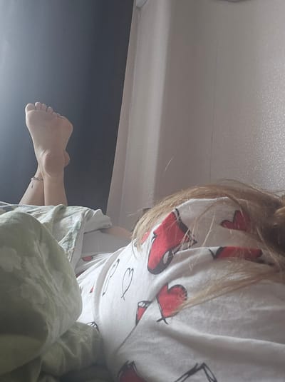 The image shows a person lying on a bed, with their feet propped up. One foot is clearly visible, with toes, the sole, and an ankle bracelet. The person is wearing a white shirt with red heart patterns. The background consists of a dark wall on the left and a light-colored textured wall on the right, possibly a curtain or wallpaper. There are also several pillows and a blanket on the bed. The lighting suggests it is daytime, with light coming from the left. The image is taken from a low angle, looking towards the person's feet and torso. There is no discernible information in the image to indicate the location in the city of Kimry, Russia.