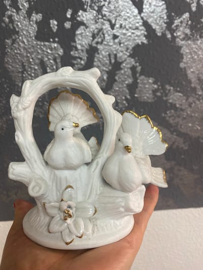 This is a close-up, eye-level, indoor photograph of a white porcelain figurine of two doves nestled on a branch with a flower at its base. The doves are white with gold accents on their beaks and tail feathers. The flower is also white with gold accents. The figurine is held in the hand of a person whose thumb and index finger are visible. The background is a textured, dark gray wall with some dappled light. The figurine is the central subject and is well-lit. The image was taken in Sachsenheim, Germany.
