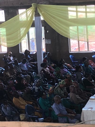 The image depicts an indoor scene, possibly a church or meeting hall, in Nteje, Nigeria. Rows of people are seated in chairs. The room is illuminated by natural light filtering through windows, some of which are adorned with pale yellow fabric drapes. A horizontal white beam or structure is visible across the top of the frame, also decorated with yellow fabric. The environment appears somewhat aged, judging by the building's structure and design.