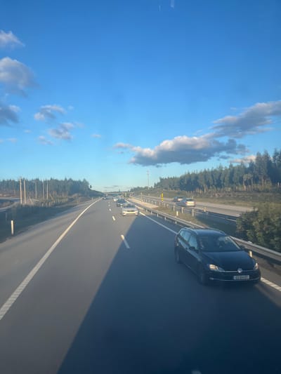 The image displays a multi-lane highway scene near the city of Pombal, Portugal, under a clear blue sky adorned with scattered white cumulus clouds. The low sun, positioned to the viewer's upper left, casts extended shadows diagonally across the asphalt pavement, suggesting either early morning or late afternoon light.
The foreground is dominated by a black Volkswagen Golf station wagon (license plate '02 XM 37') traveling in the right-most lane, moving away from the camera's perspective. Two occupants are visible within the vehicle. Ahead and to the left of this car, a queue of vehicles, including a white SUV and several sedans, occupies the adjacent lane, indicating moderate to heavy traffic or congestion extending into the distance. On the opposing carriageway, separated by a central reservation, multiple cars are also visible, traveling in the opposite direction.
The highway features distinct white lane markings, a continuous metal guardrail along the right shoulder, and utility poles along the left side. A tall, slender mast, likely for communication or monitoring, is situated within the central reservation. The environment surrounding the highway consists of dense, uniform stands of tall, slender trees, predominantly eucalyptus, lining both sides of the road.