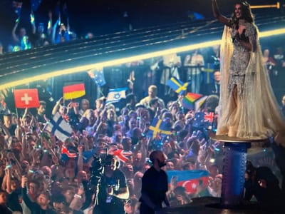 The image captures a lively scene at an outdoor concert or event, likely part of a festival, with a large crowd in the foreground and a performer on a raised stage in the background. The performer, a woman wearing a sparkling dress and a flowing cape, is holding a microphone and waving. The crowd is energetic, with many people holding up flags of various countries, including Switzerland, Germany, Sweden, Israel, Finland, and the United Kingdom. There are also rainbow flags visible. Some individuals in the crowd are holding up their phones, presumably to take photos or videos. A camera operator is present in the lower left corner of the image, near the front of the crowd. The overall atmosphere appears celebratory and engaged. The location is specified as the city of Ellwangen, Germany, though this information is not visually derivable from the image itself.