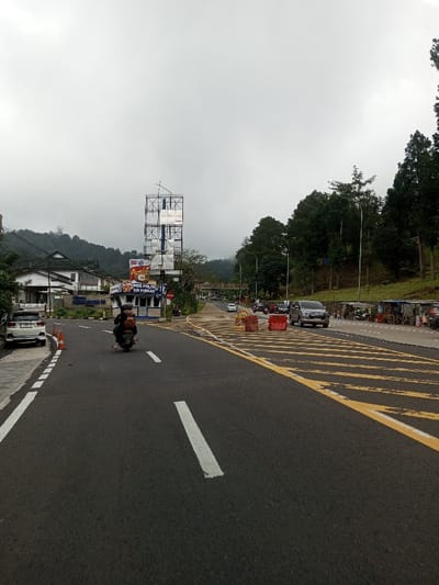The image depicts a street scene set in a hilly, possibly mountainous, region of Saing Ta Mawt Lay, Myanmar, under an overcast sky. A paved two-lane asphalt road dominates the foreground and midground, marked with white dashed lane lines and diagonal yellow stripes indicating a merge or turning area. A black motorcycle, carrying two individuals (a driver in dark attire and helmet, and a passenger with a backpack), travels away from the viewer in the left lane.
To the left of the road, a light blue and white building is identified by signage as a "POS POLISI TUR PUNCAK" (Police Post Puncak). Above this structure, a tall, multi-tiered metal framework, possibly a billboard support or communication tower, rises against the sky. Further back on the left, a white sedan is parked on the shoulder near an orange traffic cone, and other buildings are visible nestled into the hillside.
To the right of the road, dense green trees and foliage cover an upward sloping terrain. A silver SUV is visible in traffic, facing the camera, further down the road on the right, where a line of small vendor stalls and parked motorcycles also extends along the roadside. Red and yellow traffic barriers are placed on the road near the yellow diagonal markings. A pedestrian bridge spans the road in the mid-distance. Traffic appears present in both directions, indicating an active thoroughfare.