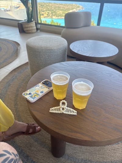 The image shows a circular wooden coffee table with two plastic cups of beer, a mobile phone with a patterned case, and a pearl hair clip. A person's feet, wearing brown sandals, are visible on the left side of the frame, next to a yellow garment. The setting appears to be a modern lounge area with large windows offering a view of the sea and a coastal landscape, likely in Agios Nikolaos, Crete, Greece, given the visual cues of a Mediterranean coastline. The background includes a plant in a woven pot, upholstered seating, and other circular tables. The overall impression is one of relaxation and leisure in a scenic location.