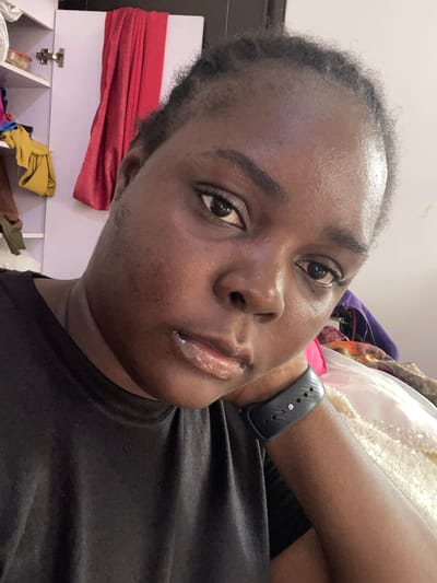 A close-up selfie shows a dark-skinned person with short, dark hair. The individual's face is slightly tilted towards the left, and their eyes are directed towards the camera. The person wears lip gloss on their lips. They are wearing a black t-shirt and a black smartwatch. The background is cluttered with items, including what appears to be a white cabinet or shelf with red fabric draped over it. Other pieces of clothing in various colors are visible in the background. The location appears to be indoors. There's no indication that this image was taken in Dushepe, Nigeria.