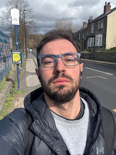 A selfie photograph features a man with a beard and blue-framed glasses. He wears a grey sweater under a black jacket and is standing at a bus stop on Talbot Street in Sheffield, United Kingdom. The Talbot Street bus stop sign is visible. Buildings are in the background across the street.