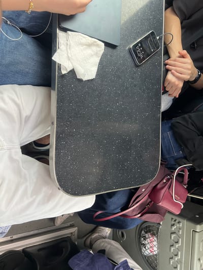 The image shows a top-down view of people sitting at a table, likely on a train or bus, given the narrow space and the presence of luggage. A black speckled table is in the center, with a laptop and a phone charging on it. There are at least three people visible, with legs and arms protruding into the frame. One person on the left is wearing blue jeans and a white top. Another person in the center is wearing white pants and grey sneakers. A third person on the right is wearing dark clothing and has their arms crossed. A dark red purse is placed on the floor next to the table. The environment appears to be indoors, with a metallic structure and luggage visible below the table. There is no information in the image to suggest the location is in Akhtala, Armenia.