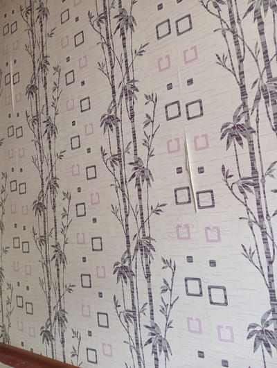 The image shows a close-up of wallpaper with a repeating pattern of bamboo stalks and leaves in shades of gray and purple. Scattered among the bamboo are small squares and larger, open squares in a light pink and dark gray color. The wallpaper appears to be slightly peeling in one section on the right side of the image. The background of the wallpaper is a textured off-white. The image is taken from an eye-level perspective, focusing on the wall's surface. There are no people or any other discernible objects or actions in the frame besides the wallpaper. The context of Gerey-Shoron, Kyrgyzstan, is not visually represented in the image itself.