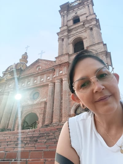 An adult female is depicted in a close-up, low-angle, selfie-style photograph positioned in front of the San Calixto Church in Timaná, Colombia. The female, in the lower right foreground, wears eyeglasses, a white sleeveless top, gold hoop earrings, and a gold chain with a small pendant. A dark band is visible on her upper right arm. She is looking towards the camera with a neutral expression.

The background is dominated by the red brick architecture of the church, which features a prominent multi-tiered bell tower with a visible clock face, a main facade characterized by large fluted columns, arched entranceways, and a domed section. Multiple crosses top the church structure against a clear, light blue sky. A series of brick steps leading to the church entrance are visible in the lower left and center. A distinct lens flare is present in the middle-left portion of the image.