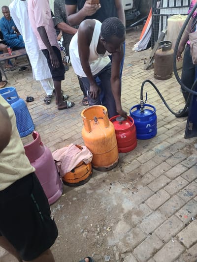 A group of men are gathered around propane gas cylinders on a paved area in Karu, Nigeria. In the foreground, a man wearing a white tank top and black shorts is leaning over, possibly attending to a red gas cylinder labeled "8.0". To his left are a large pink gas cylinder and an orange gas cylinder with a cloth draped over it. Behind these, a blue gas cylinder is connected to a hose, with a man in dark clothing and sandals standing nearby. To the left of the blue cylinder, another large blue gas cylinder is visible. Further back, men in traditional attire, including a white agbada and a pink shirt, are standing. One man in a blue tunic is seated on a low table with a soda can on it. The environment appears to be an outdoor, possibly commercial, area with a rough, paved surface and some debris. The lighting suggests it is daytime.