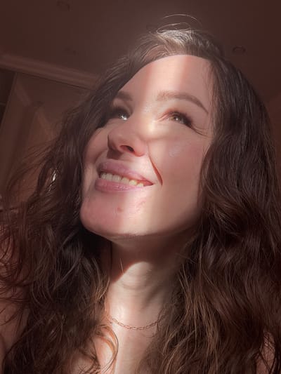 A young woman with long, wavy dark brown hair is captured in a close-up, angled portrait, likely in Yuzhny, Russia. The image is rotated, presenting her face on a diagonal tilt, suggesting she might be lying down or leaning back. A strong, warm diagonal beam of sunlight illuminates the left side of her face, highlighting her light brown or hazel eye, nose, and a portion of her smiling mouth, which reveals healthy teeth. Her eyebrows are well-defined, and she has long eyelashes.

Her expression conveys a sense of gentle happiness or contentment. Her dark hair cascades around her, partially obscuring her neck and shoulders, where a delicate gold-colored chain necklace is visible. The rest of the scene is cast in shadow, with only faint outlines of what appears to be an indoor setting, possibly a wall or furniture, in the background. The distinct sunlight suggests either an early morning or late afternoon time of day. There is no visible text in the image.