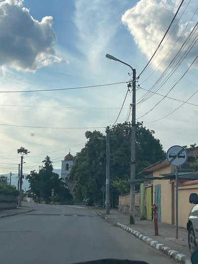 The image depicts a street scene in Novo Zhelezare, Bulgaria. The view is from a low angle, suggesting it was taken from inside a vehicle. The street is paved and appears to be relatively quiet. On the right side of the street, there is a building with a light-colored facade, a green door, and a turquoise door. A red fire hydrant stands on the sidewalk in front of this building. A traffic sign depicting two cars with a diagonal line through them, indicating the end of an overtaking restriction, is visible. There are several utility poles with numerous overhead wires crisscrossing the sky. In the background, a white church with a red-domed roof is partially visible amidst lush green trees. The sky is blue with scattered white clouds. The overall impression is of a tranquil, somewhat rural, European town setting.