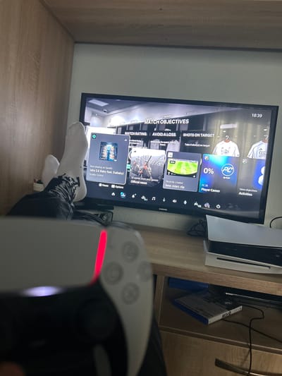The image shows a person playing the game FC 26 on a television screen using a PlayStation 5 controller. The television displays in the corner of the screen "18:39." The person is wearing socks with a black pattern. Other objects visible are a PlayStation 5 console and a game case. The environment appears to be an indoor living space with a wooden shelf. There is no information provided regarding the location in the city of Hvostiane, Bulgaria, or anything specific to identify that the event took place there.