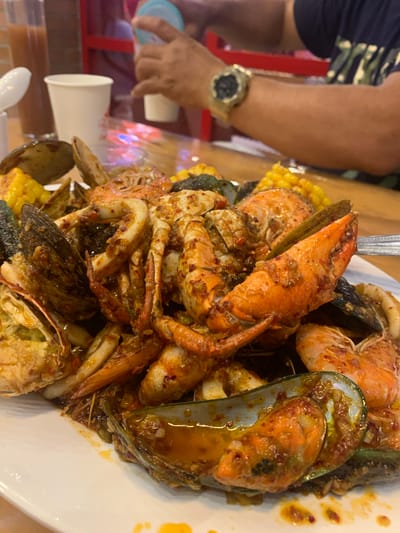 In Angeles City, Philippines, a close-up shot focuses on a large platter of mixed seafood, likely prepared in a Cajun-style sauce. The seafood includes mussels, shrimp, and pieces of crab, all coated in a glistening orange-red sauce with visible chili flakes and bits of garlic. Interspersed with the seafood are pieces of corn on the cob. The platter is positioned on a wooden table. In the blurred background, a person's hands are visible, holding a white cup and a clear cup with a blue lid, suggesting they are in the process of drinking or serving beverages. Another person's arm, wearing a watch, is also visible, indicating a social dining setting. White paper cups and a tall glass containing a brown liquid are also present on the table. The lighting suggests an indoor environment, possibly a restaurant.