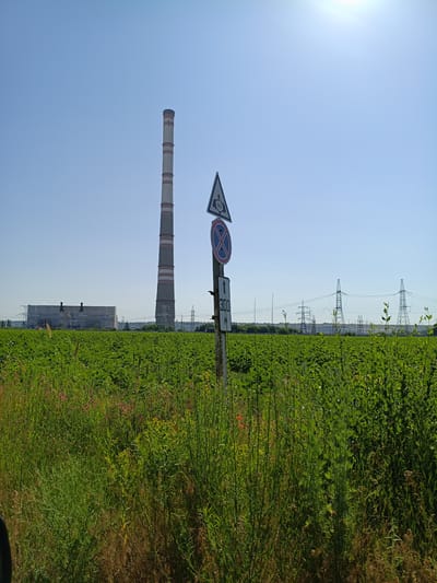 In Dnestrovsk, Moldova, a tall, cylindrical industrial smokestack with red and white bands stands in the background, dominating the skyline against a clear blue sky. To its left, a long, rectangular industrial building with a lighter-colored roof is visible. In the middle ground, a lush green field of crops stretches across the landscape, interspersed with taller grasses and some small pink and yellow wildflowers in the foreground. Several power lines and transmission towers are scattered across the horizon, suggesting an industrial or power generation area. In the foreground, a metal pole with multiple signs is present. One sign is a triangular warning sign with a symbol of a gas pipeline, another is a circular sign with a red border and a diagonal red bar over a blue background, indicating a prohibition. Below these, a rectangular sign points upwards with the number "300" and the letter "M" beneath it, likely indicating a distance. The overall scene depicts an industrial landscape bordering a rural agricultural area under a bright, sunny sky.