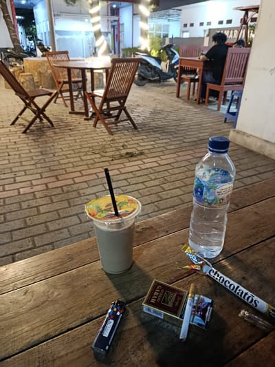 The image depicts an outdoor seating area at what appears to be a casual eatery or cafe in Sumberejo, Indonesia. The foreground features a weathered wooden table with several items placed upon it. These include a plastic cup with a straw containing a light-colored beverage, a bottle of water, a cigarette lighter with a colorful pattern, a partially opened pack of Gudang Garam cigarettes with two cigarettes, one lit, and a chocolate snack. In the background, the paved area is furnished with wooden tables and chairs. Beyond that, there are buildings with exterior lighting, and a motorcycle is visible parked to the left. A person in a black shirt and dark pants is seated at a table in the middle ground, facing away from the camera. The overall scene suggests an evening setting.