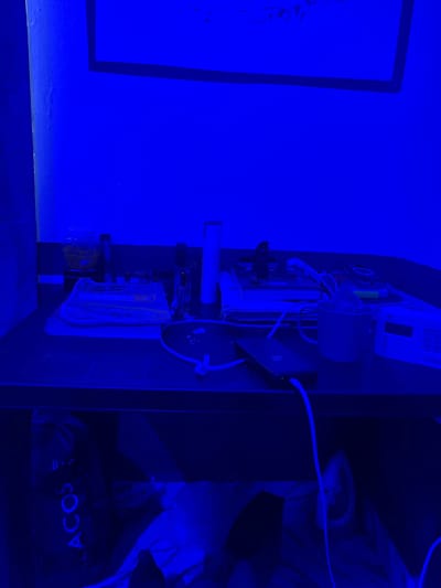 This media file shows a vertically oriented shot of a collection of electronic components and wiring, mounted on a dark surface. The scene is illuminated with a strong blue light, casting a dark blue hue over everything and obscuring many finer details. In the foreground, a blue rectangular frame is visible on the left, possibly part of a larger display or artwork.

The main subject is a cluster of electronic devices. There appears to be a cylindrical component, possibly a motor or speaker, with wires extending from it. Below this, there's a circuit board with various chips and connectors. Other components, including what look like power supplies or adapters, are also present. Some sort of container, perhaps a jar or bottle, with yellowish contents is also visible. A piece of fabric or paper with some patterned design is attached to the assembly.

In the background, to the right of the electronic components, a dark area reveals the faint outline of what could be a person wearing a light-colored garment. A dark object with the partial text "ACO" is visible near the bottom right, possibly part of a brand logo on clothing or an item. The overall atmosphere is one of technical assembly or experimentation, with the intense blue light adding a dramatic or mysterious quality. No specific time of day or weather conditions can be determined due to the artificial lighting and enclosed setting. The location context of Pantinapu, Nigeria, is not visually supported by any specific cues within the image.