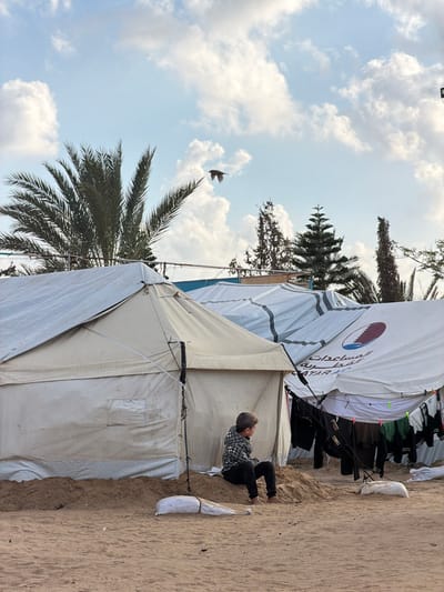 The image captures an outdoor scene in a-Zawayda, Palestinian Territory, featuring a temporary settlement under a partly cloudy sky. A young male child, approximately 5-8 years old, is seated on a low sand mound in the foreground. He wears a plaid long-sleeved shirt, dark trousers, and has bare feet. He faces right, looking away from the camera. Behind him and to his left stands a light-colored canvas tent with visible guy lines. To the right of the child, a larger tent is covered with a white tarp, featuring a prominent circular logo with maroon and blue elements, along with Arabic text "المساعدات القطرية" (Qatari Aid) and "QATAR AI..." partially visible. Clothing, including dark shirts and lighter garments, hangs on a line suspended in front of this branded tent, secured by clothespins. Several weighted white bags, likely sandbags, are positioned on the sandy ground, acting as anchors for the tents. The ground consists of light brown sand or loose earth. In the background, a large palm tree with green fronds is visible on the left, alongside several coniferous and other sparse trees behind the tent structures. The sky is light blue with scattered white cumulus clouds. A dark bird is captured in flight in the upper center of the frame.